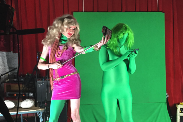 Two performers against a green screen and red curtains. One in a pink dress and blonde wig holds a selfie stick; the other in
